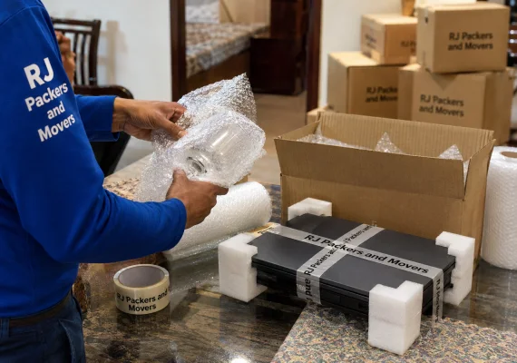 RJ Packers and Movers fragile glassware and electronics being double-protected with bubble wrap, foam, corner guards, and RJ Packers and Movers packing tape printed RJ Packers and Movers in black text only before insured transit