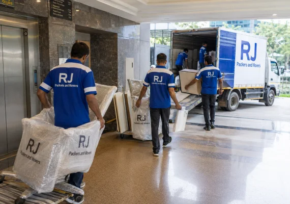 RJ Packers and Movers movers carrying wrapped office chairs and dismantled desks through a commercial building lobby toward a white-blue truck